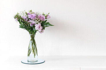 Bouquet Of Purple Lisianthus And Baby's Breath Flowers Is Standing In A Vase On A White Table Against A White Wall