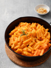 Close-up of a black bowl filled with creamy mac and cheese, garnished with a sprig of rosemary. A small white dish with breadcrumbs is blurred in the background on a gray surface.
