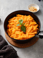Close-up of a black bowl filled with creamy mac and cheese, garnished with a sprig of rosemary. A small white dish with breadcrumbs is blurred in the background on a gray surface.
