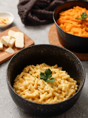 Two black bowls of macaroni and cheese on a gray surface. The front bowl contains creamy mac and cheese, garnished with herbs. In the background, a second bowl features a different cheese sauce