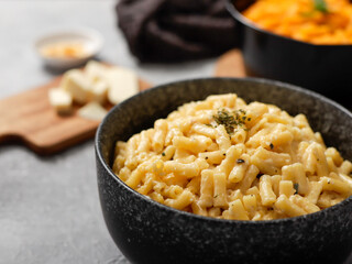 Two black bowls of macaroni and cheese on a gray surface. The front bowl contains creamy mac and cheese, garnished with herbs. In the background, a second bowl features a different cheese sauce