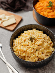 Two black bowls of macaroni and cheese on a gray surface. The front bowl contains creamy mac and cheese, garnished with herbs. In the background, a second bowl features a different cheese sauce