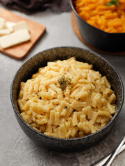 Two black bowls of macaroni and cheese on a gray surface. The front bowl contains creamy mac and cheese, garnished with herbs. In the background, a second bowl features a different cheese sauce