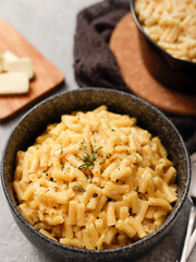 Two black bowls filled with creamy macaroni and cheese, garnished with rosemary, sit on a gray textured surface. Cheese slices on a wooden board are visible in the background a dark cloth.