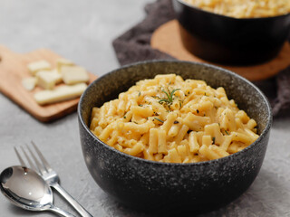 Two black bowls filled with creamy macaroni and cheese, garnished with rosemary, sit on a gray textured surface. Cheese slices on a wooden board are visible in the background a dark cloth.