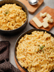 Two black bowls filled with creamy macaroni and cheese, garnished with rosemary, sit on a gray textured surface. Cheese slices on a wooden board are visible in the background a dark cloth.