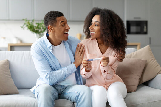 Happy African American spouses holding positive pregnancy test and looking at each other, excited of becoming parents, sitting on sofa at home