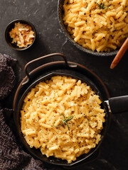 Two black bowls filled with creamy macaroni and cheese, garnished with rosemary, sit on a gray textured surface. Cheese slices on a wooden board are visible in the background a dark cloth.