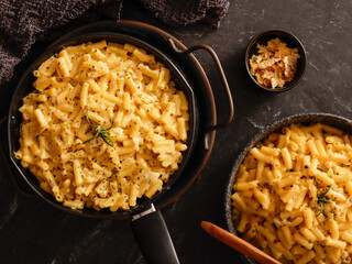 Two black bowls filled with creamy macaroni and cheese, garnished with rosemary, sit on a gray textured surface. Cheese slices on a wooden board are visible in the background a dark cloth.