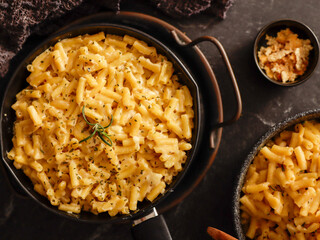 Two black bowls filled with creamy macaroni and cheese, garnished with rosemary, sit on a gray textured surface. Cheese slices on a wooden board are visible in the background a dark cloth.