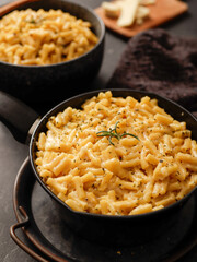 Two black bowls filled with creamy macaroni and cheese, garnished with rosemary, sit on a gray textured surface. Cheese slices on a wooden board are visible in the background a dark cloth.