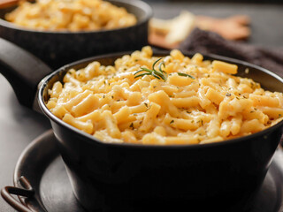 Two black bowls filled with creamy macaroni and cheese, garnished with rosemary, sit on a gray textured surface. Cheese slices on a wooden board are visible in the background a dark cloth.