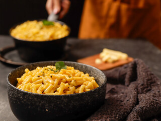 A black bowl filled with creamy macaroni and cheese, garnished with fresh basil leaves, sits on a dark surface. A blurred hand in the background is holding another bowl of the same dish.