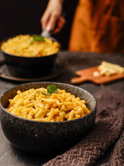 A black bowl filled with creamy macaroni and cheese, garnished with fresh basil leaves, sits on a dark surface. A blurred hand in the background is holding another bowl of the same dish.