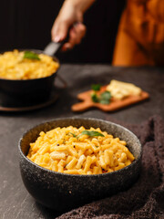 A black bowl filled with creamy macaroni and cheese, garnished with fresh basil leaves, sits on a dark surface. A blurred hand in the background is holding another bowl of the same dish.