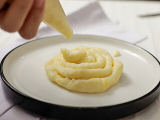 Close-up of a hand squeezing mashed potatoes from a piping bag onto a white plate. The creamy...