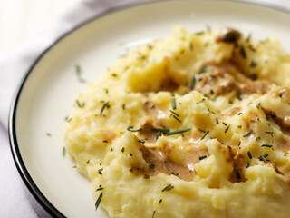 A plate of creamy mashed potatoes topped with a rich, savory gravy and garnished with fresh herbs. The dish is served on a white plate, with a spoon placed on the side, ready to be enjoyed