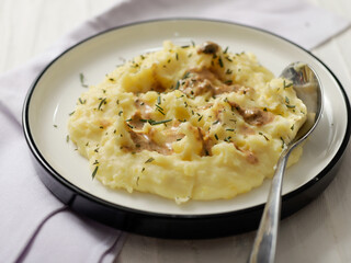 A plate of creamy mashed potatoes topped with a rich, savory gravy and garnished with fresh herbs. The dish is served on a white plate, with a spoon placed on the side, ready to be enjoyed