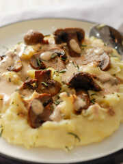 A plate of creamy mashed potatoes topped with a rich mushroom gravy and garnished with fresh rosemary. The dish is elegantly presented on a white plate, highlighting the hearty, comforting meal.