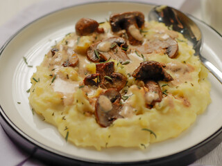 A plate of creamy mashed potatoes topped with a rich mushroom gravy and garnished with fresh rosemary. The dish is elegantly presented on a white plate, highlighting the hearty, comforting meal.