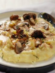 A plate of creamy mashed potatoes topped with a rich mushroom gravy and garnished with fresh rosemary. The dish is elegantly presented on a white plate, highlighting the hearty, comforting meal.