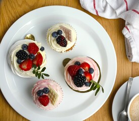 Cupcakes and mini pavlova cakes decorated with fresh berries and white chocolate on a large white plate, a cup of coffee and a cloth napkin on wooden table. Summer fruit cakes flat lay