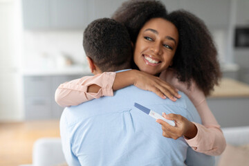 Happy black woman holding positive pregnancy test and hugging her husband, family waiting for baby, celebrating becoming parents