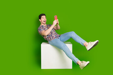 Full size photo of overjoyed guy dressed print shirt jeans sit on white cube look at smartphone...