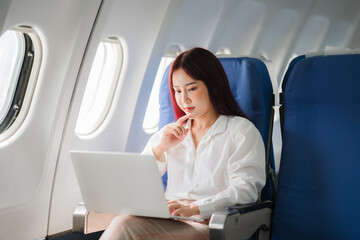 Focused in Flight: A young Asian businesswoman, bathed in the soft glow of the airplane window, contemplates her next move on her laptop, embodying the modern work-life blend. 