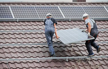 Men technicians carrying photovoltaic solar moduls on roof of house. Electricians in helmets installing solar panel system outdoors. Concept of alternative and renewable energy.