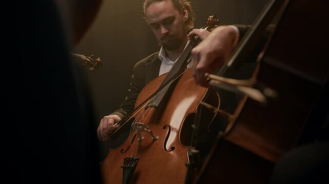 Medium shot of Caucasian male and female string quartet playing violins, cello and bass while sitting in circle under projector light on black background