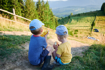 Two young boys sit on dirt path, enjoying snacks. One boy in blue shirt eats corn on cob, while other, holds bun. Back view of children overlook scenic valley with rolling hills and pine trees.