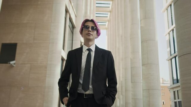 Tracking medium slowmo of cool young Asian guy with pink hair wearing elegant black suit and tie and trendy glasses walking confidently down street at daytime