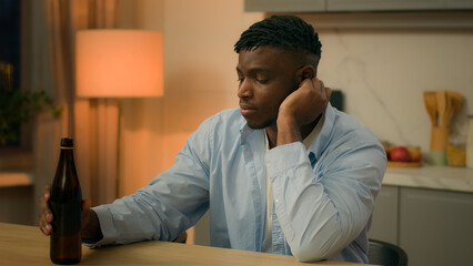 Sad upset African American man alone thinking at table in kitchen night drunk male addicted guy...