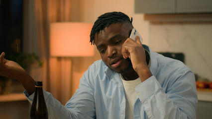 Drunk alcoholism addicted African American man guy male drinking alcohol drink beer at home evening talking mobile phone call smartphone talk speak suffering problem trouble sad upset stress depress