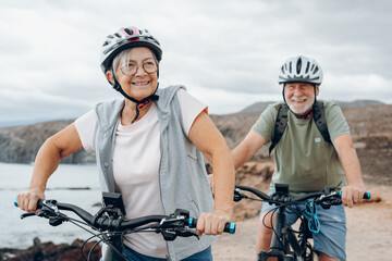 Senior couple riding bikes together in rocky beach enjoying outdoor. Active mature people talking holding mountain bikes. Bicycle lifestyle concept, having fun.