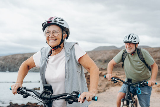 Senior couple riding bikes together in rocky beach enjoying outdoor. Active mature people talking holding mountain bikes. Bicycle lifestyle concept, having fun. - Powered by Adobe
