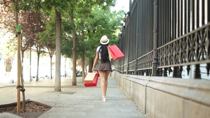 Woman with long hair and hat walking down a tree-lined sidewalk, carrying shopping bags.