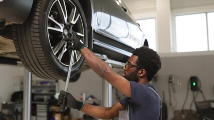 Smiling mechanic using wrench to repair car wheel in modern garage. Professional technician working on vehicle maintenance. Handsome young man wearing glasses engaging in automotive repair. - Powered by Adobe