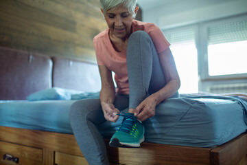 Active senior woman tying sneakers in bedroom at home