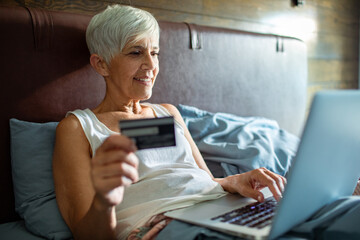Smiling senior woman using credit card on laptop in bed