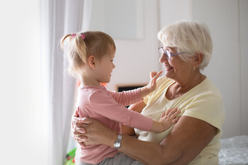 Happy loving granddaughter child hugging excited blonde grandma from behind at home. Grandmother and kid girl enjoying funny leisure on family meeting, having fun, laughing