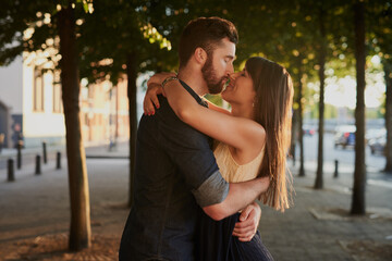 Kissing, care and couple with hug in nature for romance, connection and happiness together. Love, embracing and young people with bonding for sweet moment, commitment or marriage outdoor in city.