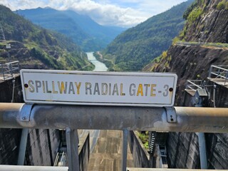 A close-up of a sign that reads "Spillway Radial Gate 3" mounted on a metal railing. The sign is positioned in front of a massive concrete dam and reservoir. 