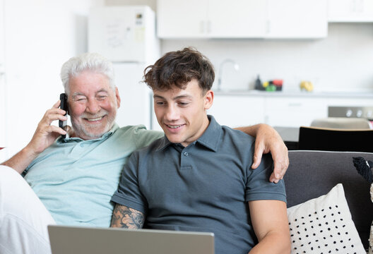 Smiling grandfather talking on cell phone sitting next to young grandson while using laptop, handsome smiling boy teaching how to use computer, how to shop online, book travel and vacation