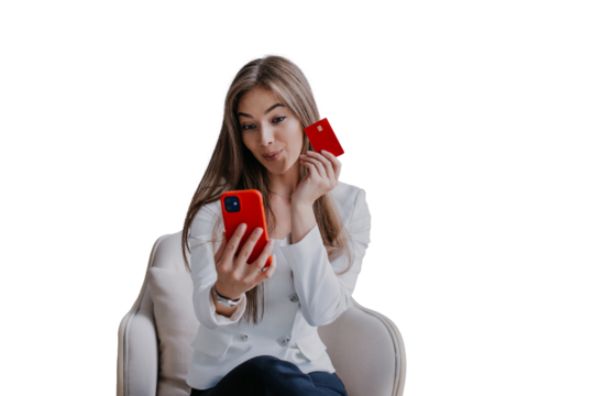 young woman sitting on a chair, holding a red smartphone and a credit card against transparent background, making an online purchase, with playful expression. Concept of online shopping 