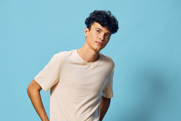 Young man with curly hair wearing a light beige t shirt against a solid blue background, exhibiting...