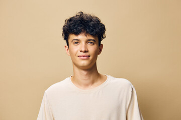 Young man with curly hair smiling softly, wearing a light colored t shirt against a warm beige background, showcasing a friendly and approachable demeanor for diverse lifestyle concepts