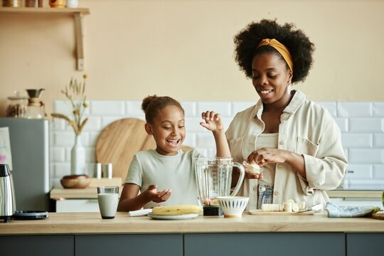 Smiling African American girl and young mother making banana smoothie on large counter having fun and sharing happy moments at cozy beige kitchen, copy space