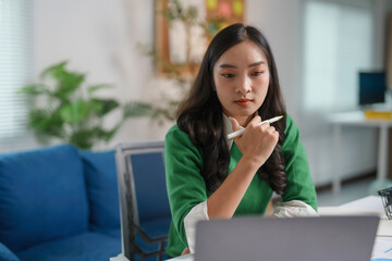 Young businesswoman is concentrating while working from home on her laptop, holding a pen to her chin in thought